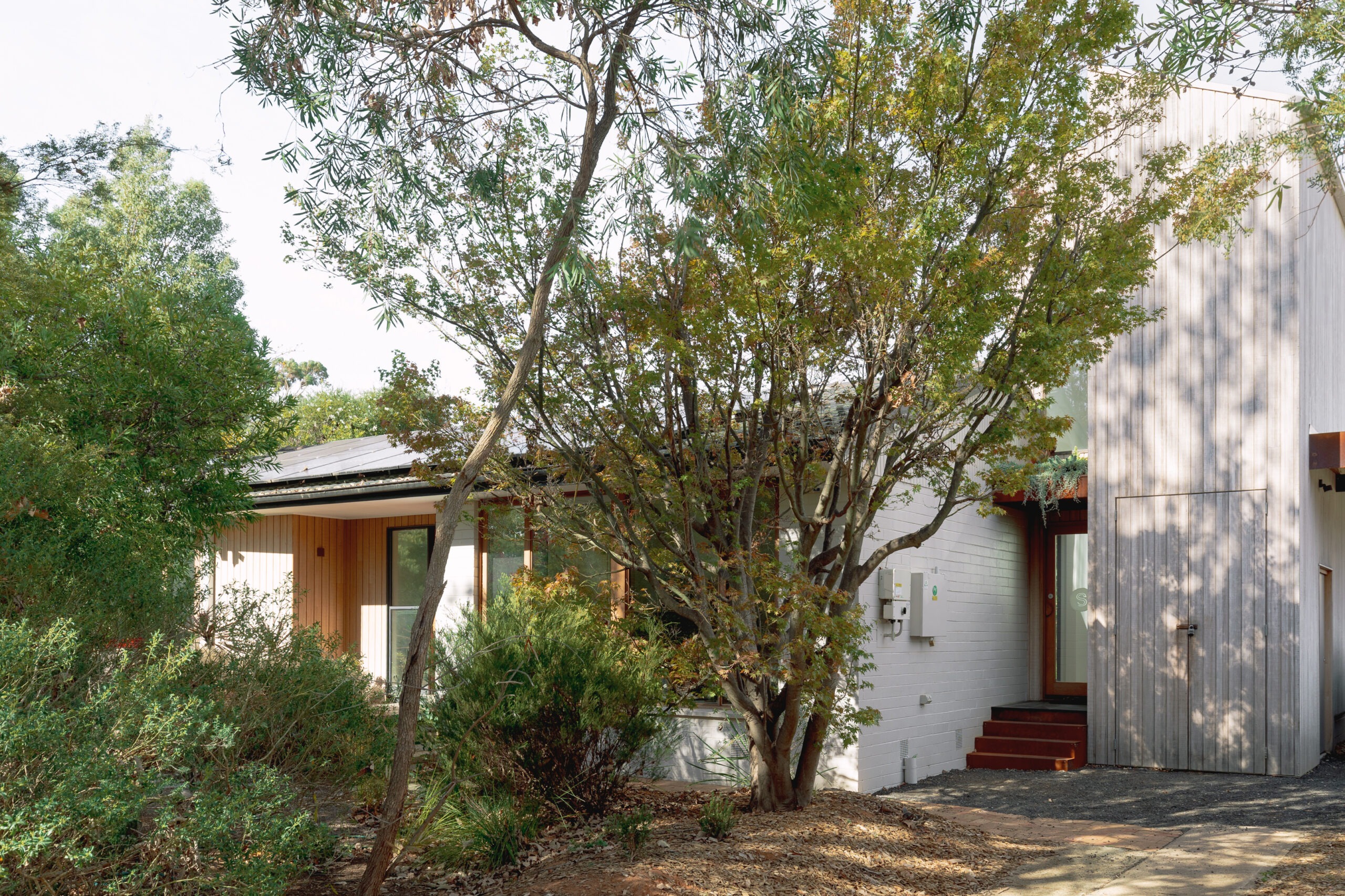 Image shows the exterior of a house in Watson, Canberra. The house is white with timber features and has a bushy garden and a small, timber clad two storey extension.